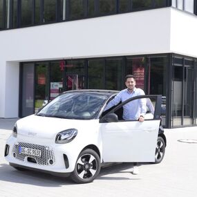 Ein Mann steht vor einem modernen Bürogebäude neben einem weißen Elektroauto Smart.
