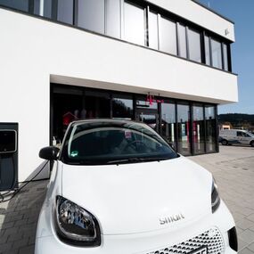 Weißes Elektroauto lädt an moderner Ladestation vor Bürogebäude bei Sonnenschein.